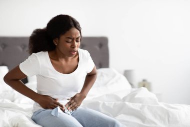 Genç siyahi bir kadın yatağında oturmuş, karnını ağrıtıyor. Pijama giyiyor ve parlak, dağınık bir yatak odasında regl kramplarıyla uğraşıyor..