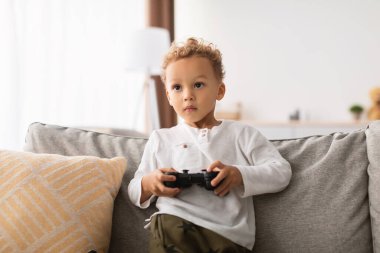 Afrikalı küçük çocuk elinde bir oyun kumandası tutarak evdeki kanepede oturuyor. O odaklanmış ve hafta sonu boyunca video oyunları oynayarak eğleniyor. Bu sahne eğlenceli bir anı yakalıyor..