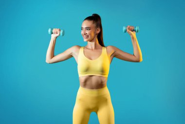 Mutlu kadın antreman sırasında dambılları tutarken kollarını esnetir. Parlak sarı fitness kıyafeti giyiyor, parlak mavi arka plana karşı güç ve coşku sergiliyor..