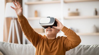 Neşeli, kilolu bir genç oturma odasındaki bir kanepede oturur, VR gözlük takar ve görünmez bir şeye dokunmak için uzanır. Modern teknolojiyle rahat bir ortamda eğleniyorlar..