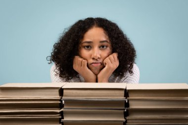 Yorgun genç siyah bir kız öğrenci büyük kitap yığınlarıyla bir masada oturuyor. O, sınava hazırlanırken sıkılmış görünüyor ve çenesini ellerinin üzerinde dinlendiriyor..