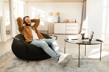 Bir adam rahat bir şekilde koltukta oturuyor, kulaklık takıyor ve gülümsüyor. Parlak bir odada dizüstü bilgisayar ve kahve masasıyla rahat bir atmosfer yaratıyor..