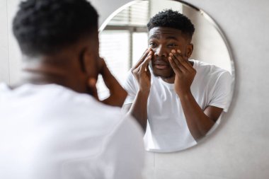 Modern bir banyoda, uykulu bir siyah milenyum adamı aynadaki yansımasına bakarken gözlerinin altına dokunur. Sabah cilt bakımının bir parçası..