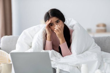 Genç bir Hintli kadın battaniyeye sarılı bir kanepede oturuyor, grip yüzünden kendini kötü hissediyor. Etrafı rahat bir oturma odasıyla çevrili dizüstü bilgisayarına bakarken burnunu siler..