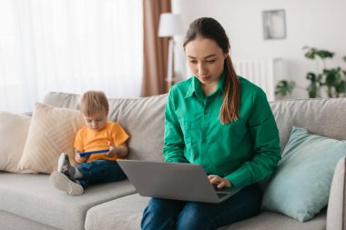 Bir anne kanepede oturur, dizüstü bilgisayarına odaklanır, genç oğlu ise yakınlardaki bir akıllı telefonda oynar. Odanın yumuşak ışıklandırması ve rahat bir atmosferi var..