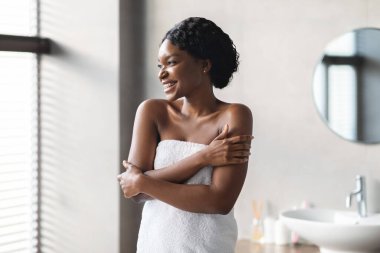 Şık bir banyoda, genç bir Afrikalı Amerikalı kadın beyaz bir havluya sarılmış bir gülümsemeyle poz veriyor. Huzurlu alana bakarken kendini kucaklıyor, kendini koruma rutinini kutluyor..