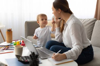Küçük bir çocuk, anneleri evde çalışırken onunla şakalaşarak etkileşime girer. Oturma odası parlak ve davetkar. Masaya serpilmiş bir sürü sanat malzemesi var..