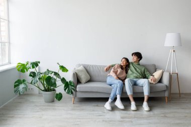 Neşeli bir Asyalı çift haftasonunu kanepede dinlenerek geçirir. Kahkahaları paylaşıyorlar ve doğal ışıkla dolu oturma odalarında birbirlerine eşlik etmekten zevk alıyorlar..