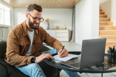 Genç bir adam modern bir oturma odasında rahatça oturup ders çalışmaya odaklanmış. Dizüstü bilgisayarını kullanarak parlak ve havadar bir atmosferle çevrili notlar yazıyor..