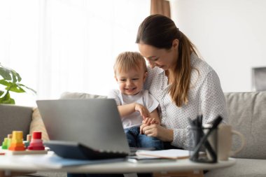 Bir anne evdeki neşeli oğluyla etkileşime girerken dizüstü bilgisayardaki işleri yönetir. Renkli oyuncaklar yakınlarda, canlı bir atmosfer yaratıyorlar..