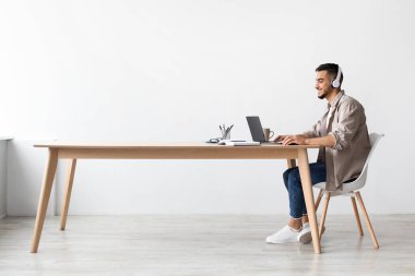 Kulaklıklı neşeli Orta Doğulu adam bir masada oturuyor ve yeni bir proje için araştırma yapıyor. Parlak, modern bir evde müzik dinlerken uzak işinden zevk alıyor..