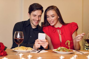 Mutlu bir çift makarnayla bir masada oturup akıllı telefona bakarken kahkahalarını paylaşıyor. Restoranın ortamı sıcak ve davetkar, mum ve şarapla dolu..