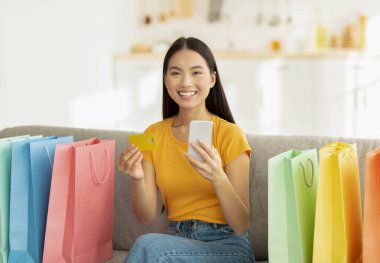 Pozitif Asyalı kadın canlı alışveriş torbalarıyla çevrili bir kanepede oturuyor. Çevrimiçi alışveriş yapmak için akıllı telefonu ve kredi kartını kullanırken kameraya gülümsüyor..