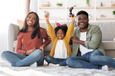 Bir aile rahat bir oturma odasında rahat bir zeminde oturur. Çocuk heyecan içinde kollarını kaldırıyor ve elinde bir oyun kumandası tutuyor..