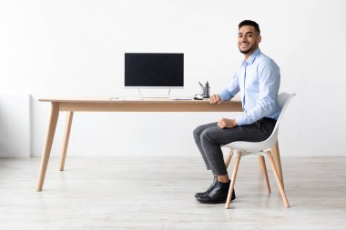 Mutlu bir Arap şık bir ev ofisinde masa başında oturur. Boş bir ekranla dizüstü bilgisayarını gösterirken gülümsüyor. Parlak alan uzak bir çalışma atmosferini yansıtıyor..