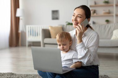 Bir anne, çocuğu yanında otururken bir telefon görüşmesi yaparken gülümser. Konforlu bir oturma odasındalar, uzak bir işin ortasında aile yaşantısını vurguluyorlar..