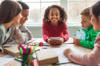 Parlak bir sınıfta, neşeli bir Afrikalı kız sınıf arkadaşlarıyla otururken telefonuna gülümsüyor. Öğrenim materyalleriyle çevrili bir okul günü birbirleriyle çatışırlar..