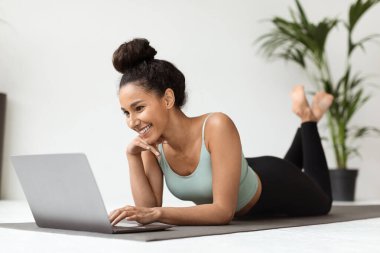 Genç bayan evde yoga minderinde vakit geçirmeyi seviyor, bilgisayarını kullanarak online fitness derslerini keşfediyor. Yeşilliklerle çevrili egzersiz rutini için video dersleri seçerken gülümsüyor..