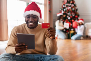 Kırmızı Noel Baba şapkalı mutlu siyah adam online Noel alışverişi yapıyor. Tablet ve kredi kartıyla. Ailesi bir Noel ağacını arkasında yumuşak bir odak noktasıyla süslüyor ve şenlikli bir ortam yaratıyor..