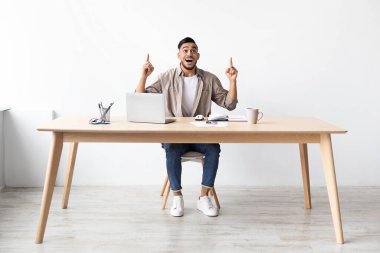 Sakallı neşeli genç bir adam parlak bir ofiste bir masada oturuyor, heyecanla yukarıya doğru işaret ediyor. Bir bilgisayar kullanıyor ve bir soruna çözüm bulmuş gibi görünüyor..