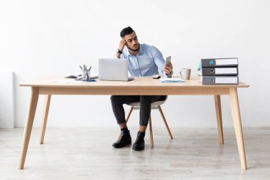 Masadaki yorgun genç adam haber okumak için cep telefonunu kullanıyor, mesai saatlerinde yorgunluk belirtileri gösteriyor. Görevlere ara verirken dikkati dağınık ve stresli görünüyor..