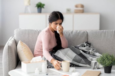 Bin yıllık Hintli kadın bir kanepede oturur, ekose bir battaniyeye sarınmıştır. Burnunu siliyor ve karantina sırasında evde soğuk belirtileriyle uğraşıyor gibi görünüyor..