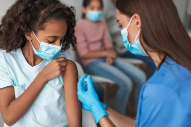 Hemşire modern bir klinik ortamında siyahi bir ergen kıza Covid-19 aşısı yapıyor. Kız maske takarken odaklanmış görünüyor. Arka planda maskeli çocuklar bekliyor..