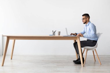 Arap bir adam ofisinde modern bir masada oturur, bir Webinar izlemek için dizüstü bilgisayar kullanır. Odaklanmış ve ciddi görünüyor, mesai saatlerinde öğrendiği gibi not alıyor..