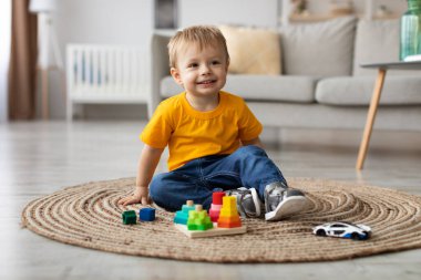 Küçük bir çocuk yuvarlak bir halıya oturur ve renkli yapı taşlarıyla mutlu mesut oynar. Konforlu oturma odasında yumuşak mobilyalar ve pencereden süzülen doğal ışık var..