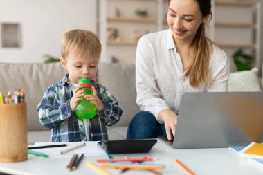Genç bir çocuk, dizüstü bilgisayarına odaklanmış olan annesinin yanında dururken yeşil bir içecek tutuyor. Konforlu oturma odasında masanın üzerine serpiştirilmiş sanat malzemeleri var..