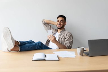 Bir Arap, sandalyesine yaslanıp ayaklarını masaya dayayarak gülümsüyor. Cep telefonunu kontrol ediyor, boş zamanlarında iş yerine ara veriyor..