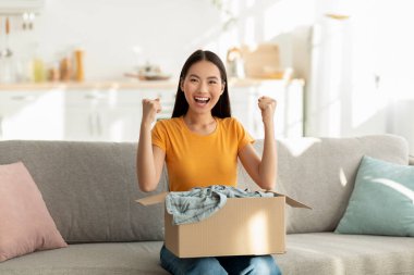 Çok mutlu bir kadın oturma odasındaki koltukta oturmuş, karton bir paketi açıyor. Mutluluktan yumruklarını sıkıyor, başarılı bir alışveriş anının tadını çıkarıyor..