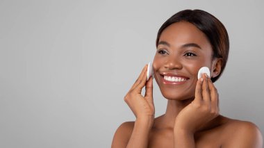 Genç bir kadın yüzüne pamuk pedlerle cilt bakımı yapıyor. Işıklı bir alanda mutlu bir şekilde gülümsüyor. Açık gri bir arka planı var. Parlayan derisini gösteriyor..