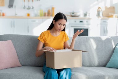 Üzgün bir bayan, parlak bir oturma odasında gri bir kanepede oturuyor ve boş bir kutuya bakıyor. Yanlış paketi almaktan duyduğu hayal kırıklığını gösteriyor. Kopyalama alanı mevcut.