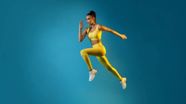 Motive olmuş genç bayan, mavi bir stüdyo arkaplanının üzerinde poz verirken dinamik enerji sergiliyor. Parlak sarı spor kıyafetleri giyiyor ve odaklanmış ve kararlı görünüyor..