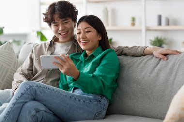 Çift evdeki rahat bir koltukta oturup gülümsüyor ve bir akıllı telefonu paylaşıyor. Rahat bir atmosferde dinlenirken mobil içeriğe dalmış bir şekilde hafta sonunun tadını çıkarıyorlar..