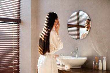 Beyaz bornozlu uzun saçlı genç Japon bayan aynadaki yansımasına gülümsüyor. Yüzüne nazikçe dokunur ve şık bir banyo ortamında hidratlaştırıcı makyaj yapar..