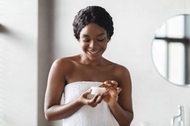 Beyaz bir havluya sarılı gülümseyen genç siyah bir kadın modern bir banyoya cilt yumuşatıcı sürüyor. Sıcak ışık, ferahlatıcı bir sabah duşundan sonra onun mutluluğunu vurgular..