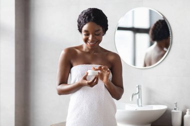 Çekici genç siyahi kadın, beyaz havluya sarılı bir vücut bakım ürünü kullanırken gülümsüyor. Sabah duşundan sonra modern banyo ortamında cilt bakımını sever..