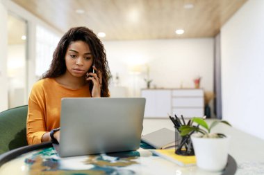 Kıvırcık saçlı bir kadın, modern bir masada bilgisayarını kullanırken bir telefon görüşmesi yapar. Çalışma alanı parlak ve düzenli, verimli bir atmosfer yaratıyor..