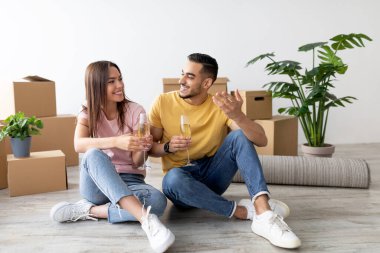 Genç bir çift yerde oturup yeni evlerinin tadını çıkarırken ellerinde içkileriyle gülümsüyor. Odada paketleri açılmış kutular ve yemyeşil bitkiler bulunuyor. Sıcak bir atmosfer yaratıyorlar..