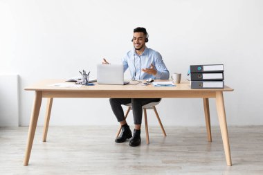 Arap bir adam ev ofisinin masasında oturuyor ve bilgisayarında bir video görüşmesi yapıyor. Konuşurken kulaklık takıyor ve gülümsüyor..