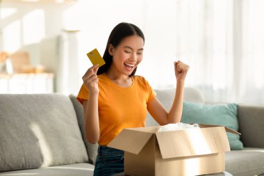 Aşırı mutlu bir kadın kanepesinde bir teslimat kutusu açar ve satın alımını evet jesti ile kutlar ve elinde bir kredi kartı tutardı. Çevrimiçi alışveriş deneyimi onu heyecanlandırdı..