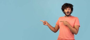 Kıvırcık saçlı ve sakallı, şaşkın Hintli bir adam pembe tişörtle yalnız poz veriyor. Mavi stüdyo arkaplanındaki boşluğu işaret ediyor. Her türlü reklam için davetkar bir ortam yaratıyor..