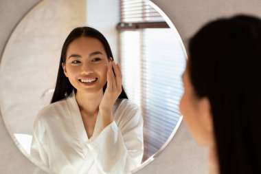 İpek bornozlu genç bir Koreli kadın, nemlendirici yüz kremi sürerken aynadaki yansımasına gülümsüyor. Parlak bir odada sabah cilt bakımının keyfini çıkarıyor..