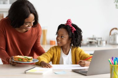 Bir anne, mutfak masasında çizim yapmaya odaklanmış kızına bir sandviç sunar. Sıcak atmosfer, öğle yemeği saatinde evdeki bağlantılarının ve yaratıcılıklarının altını çiziyor..