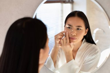 Bin yıllık Çinli bir kadın dikkatle aynaya bakıyor, yüzüne dokunuyor ve bir sivilce bastırıyor. Modern bir banyoda üzgün görünüyor, günlük hayatın baskısını yansıtıyor..