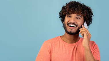 Kıvırcık saçlı neşeli bir Hintlinin çekici bir telefon konuşması yapması. Mavi bir arka plana bakarken heyecanlı görünüyor, iyi haberleri paylaşıyor..