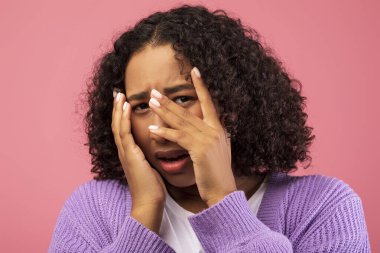 Pembe bir stüdyo ortamında, genç siyah bir kadın görünür bir şekilde korkmuş ve yüzünü panikle örtüyor. Şiddetli korku belirtileri gösteriyor. Korku ve anksiyete deneyimlerini yansıtıyor..