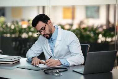 Çağdaş bir ofiste masa başında oturan bir doktor, bitkilerle çevriliyken notlar yazmaya odaklanmış. Masada bir laptop ve tıbbi ekipman var..